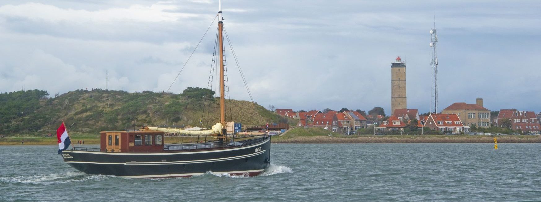 De IJver op het water, met bebouwing en een vuurtoren aan de kust op de achtergrond.